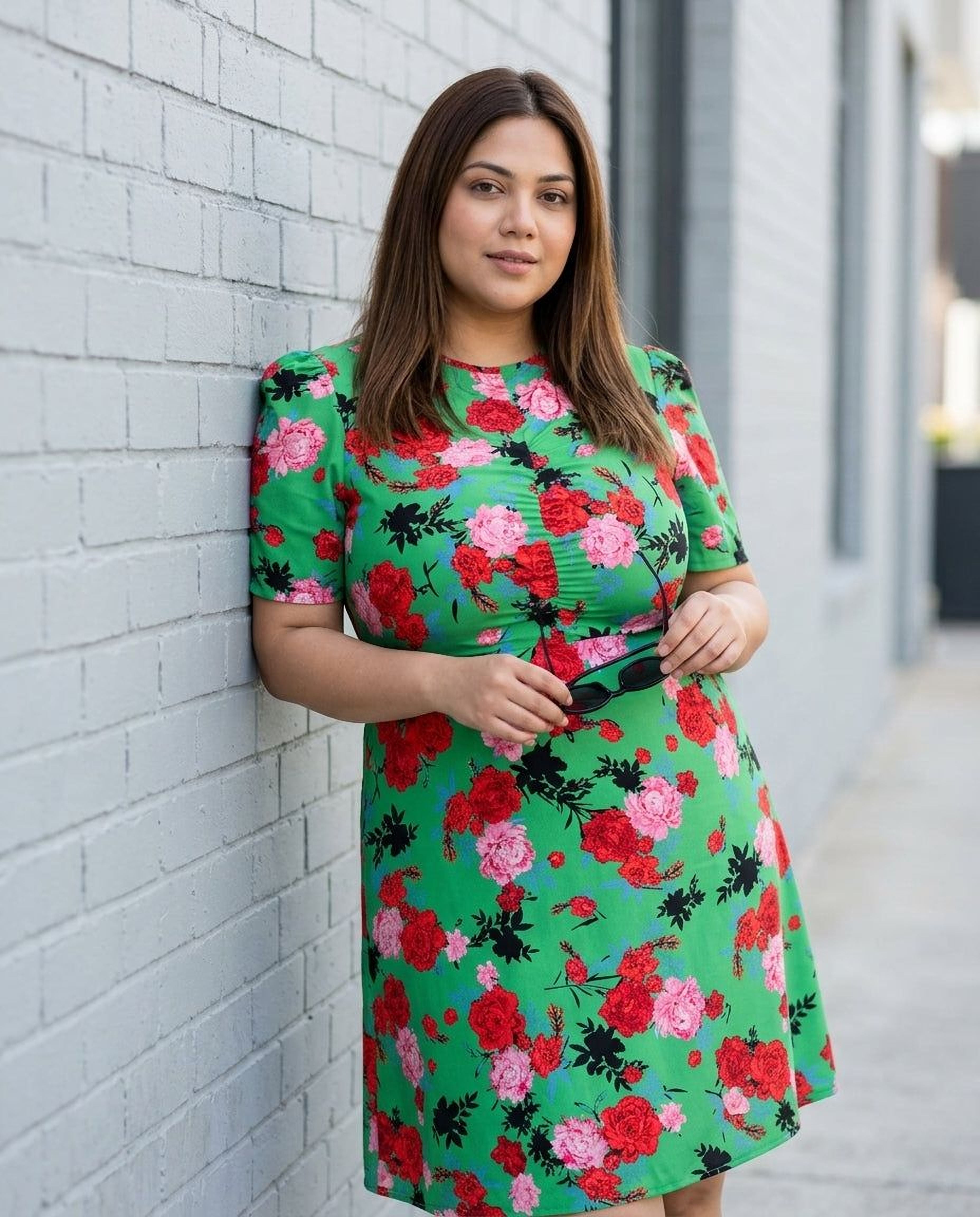 A woman wearing a green floral print dress and sunglasses, holding a black purse, and smiling at the camera.