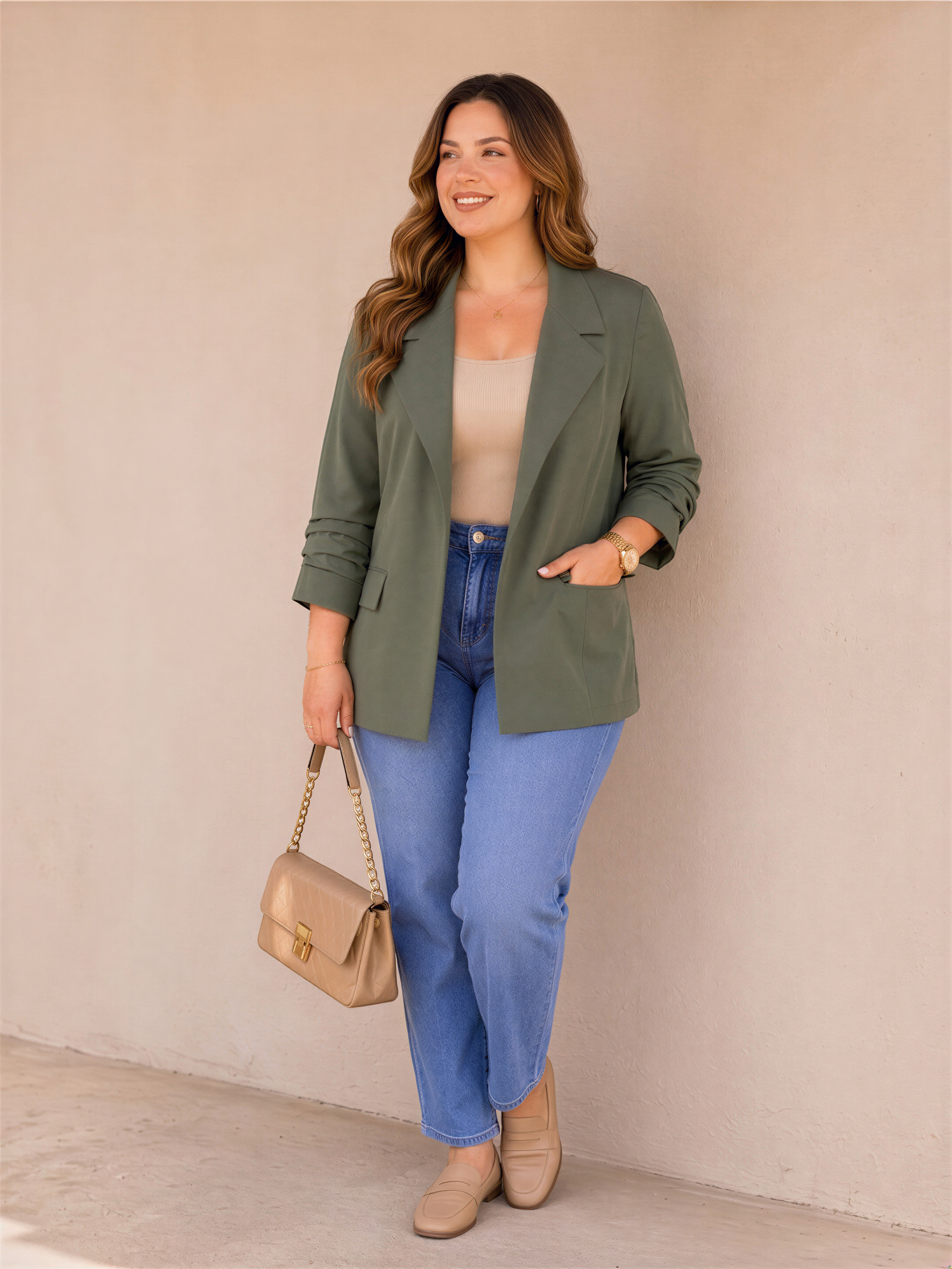 A woman in casual attire stands against a beige wall, smiling and holding a small beige handbag.