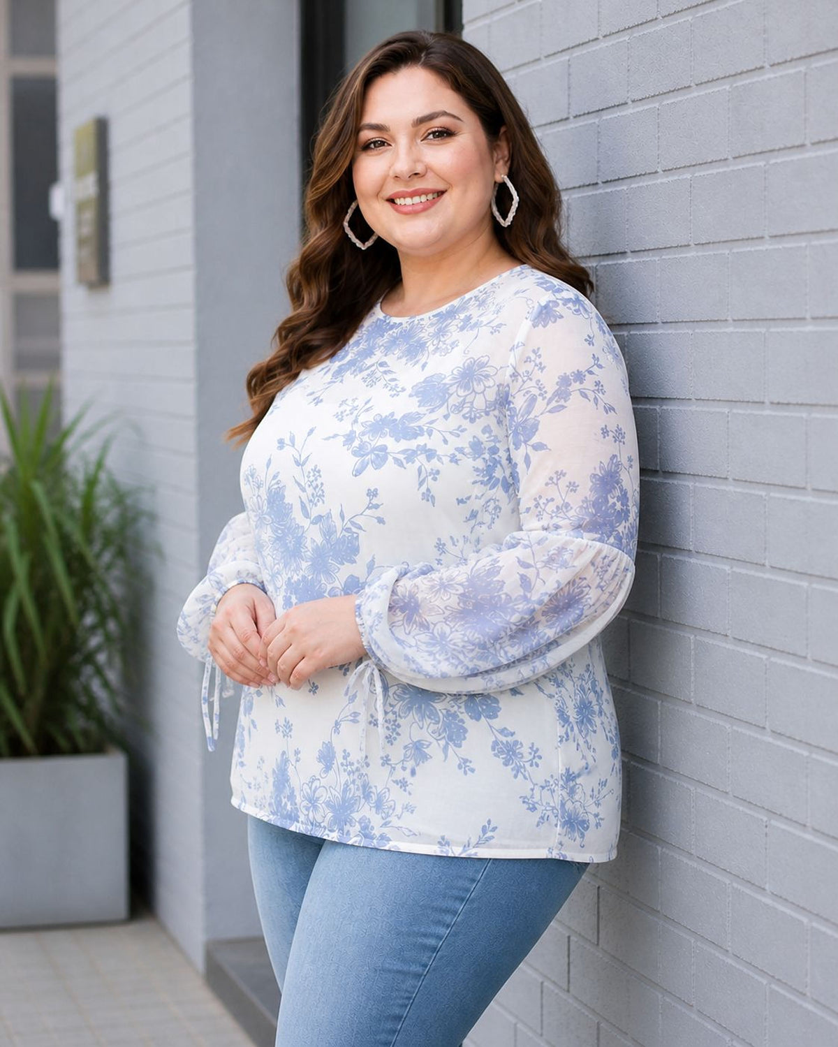 A woman with long brown hair is standing in front of a gray brick wall, smiling and wearing a white blouse with blue floral patterns.