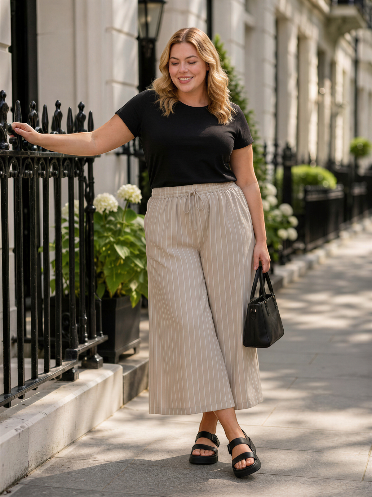 A woman in a black top and beige striped pants is leaning against a black wrought iron fence, smiling and holding a black handbag.