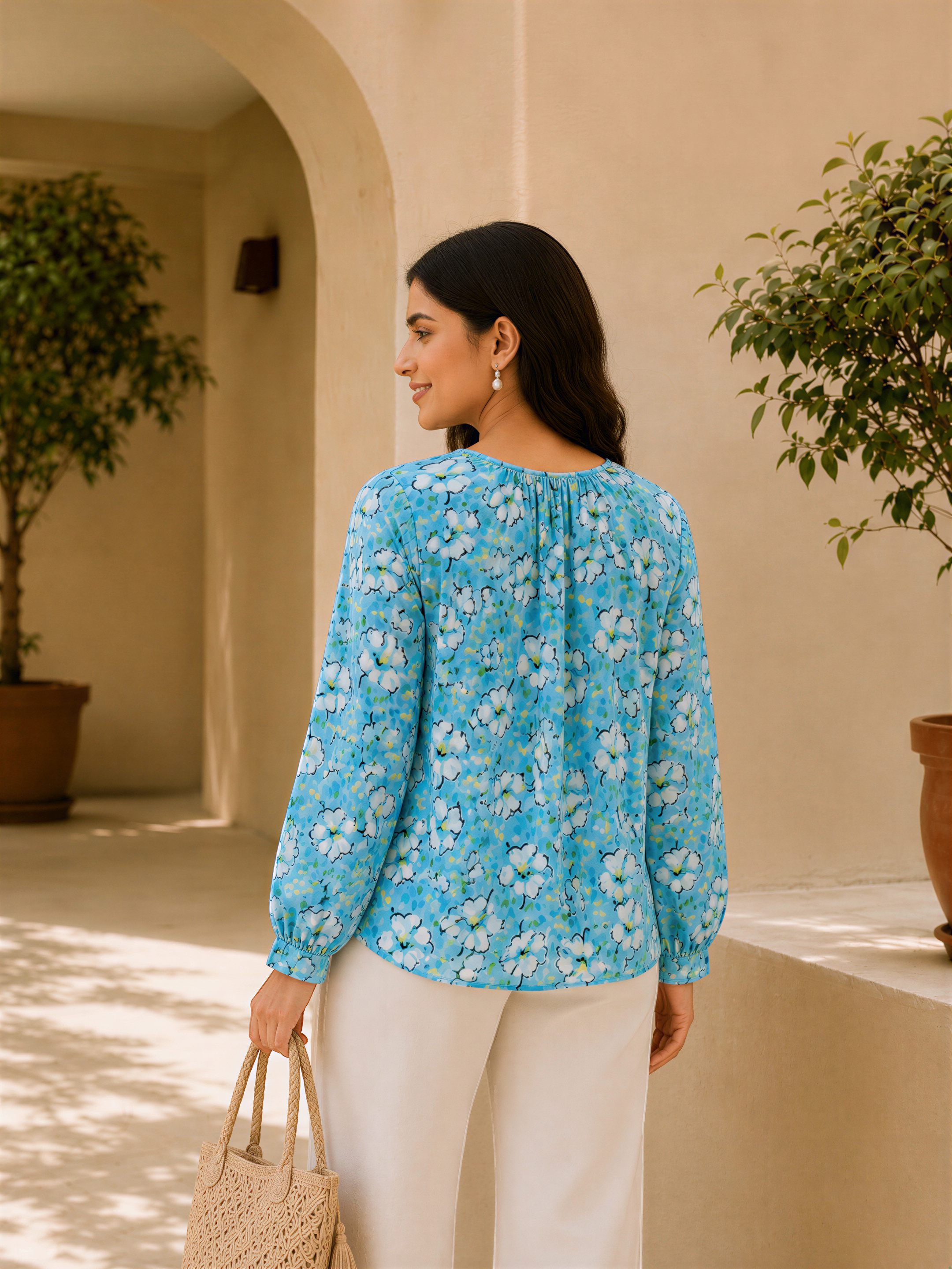 A woman wearing a blue floral blouse and white pants is standing in front of a beige wall with potted plants. She is holding a beige purse and smiling at the camera.