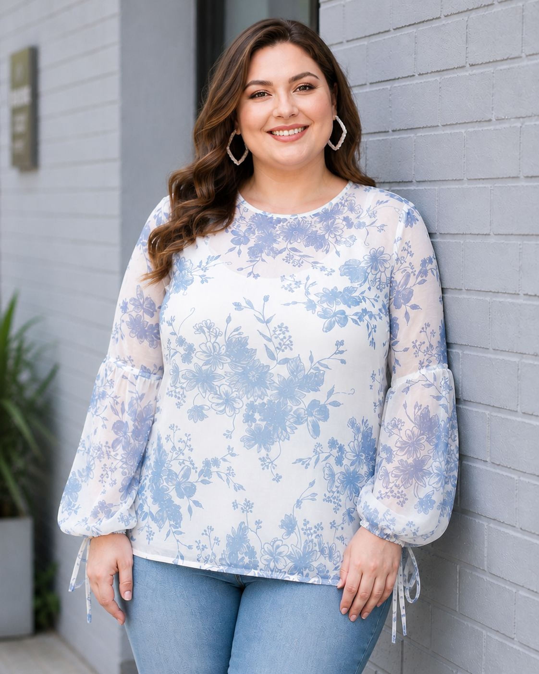 A woman with long brown hair is smiling and posing in front of a gray brick wall. She is wearing a white blouse with blue floral patterns and blue jeans.