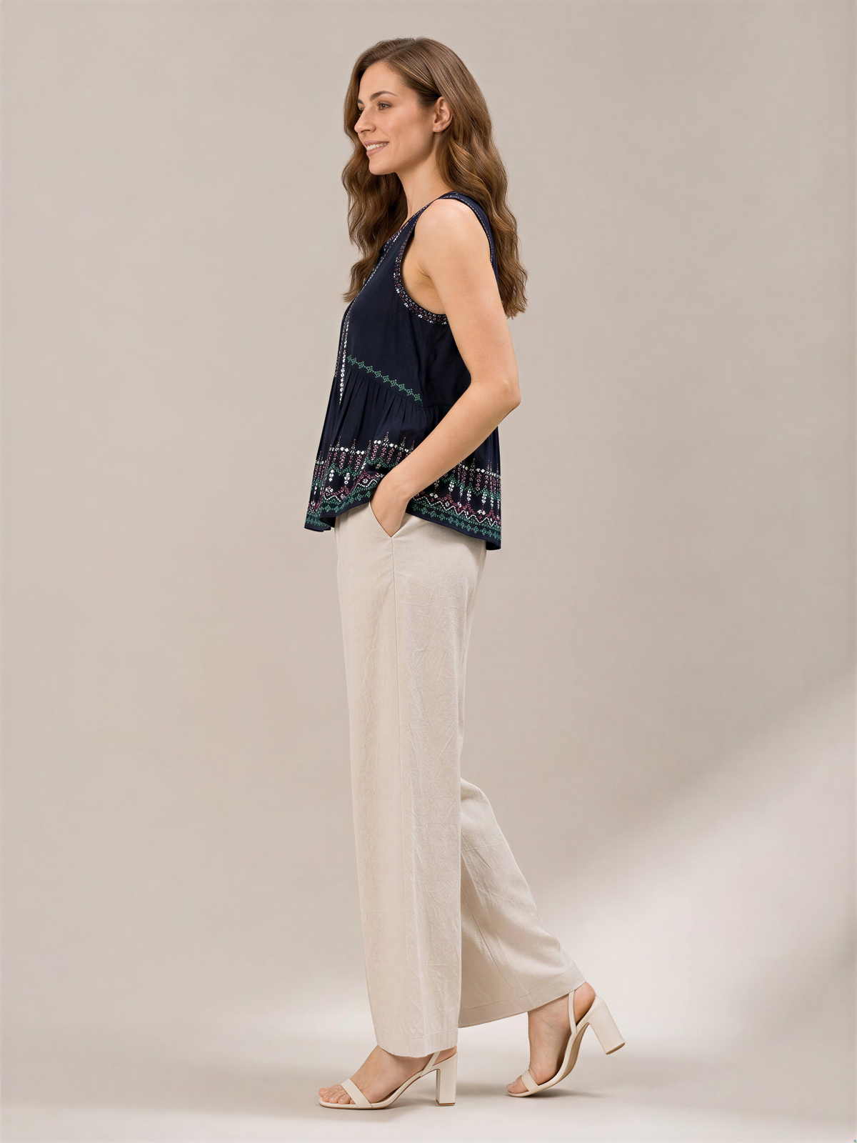 A woman wearing a navy blue top and beige pants stands against a plain background, smiling and looking to her left.