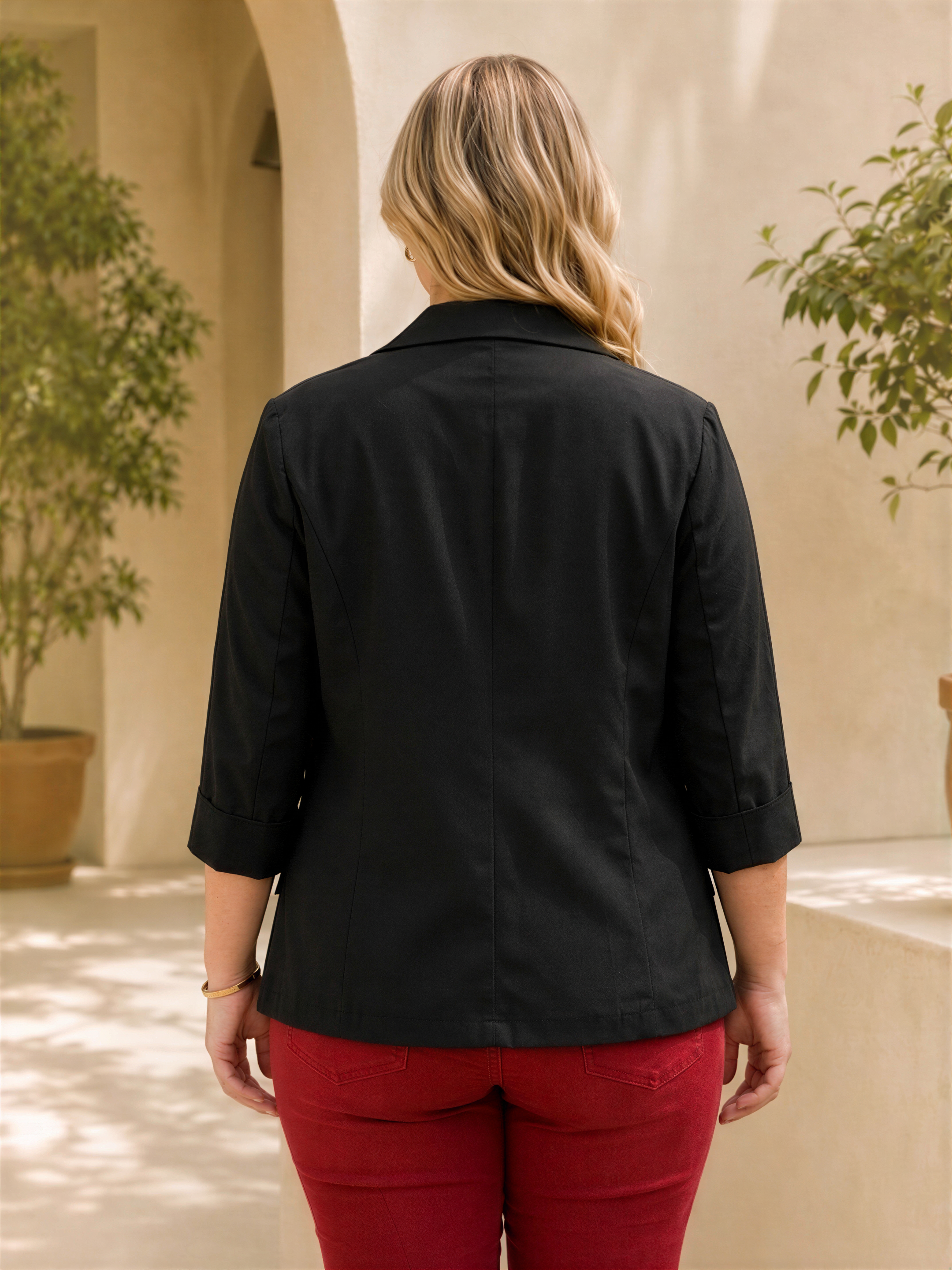 A woman wearing a black blazer and red pants stands facing away from the camera, with a potted plant and arched doorway in the background.