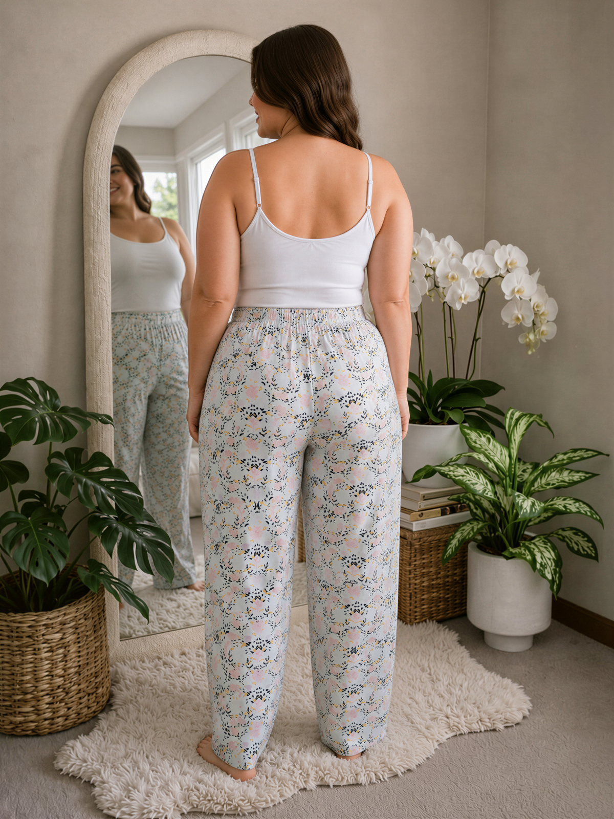 A woman stands in front of a mirror, wearing a white tank top and patterned pants, with a potted plant and a basket of books nearby.