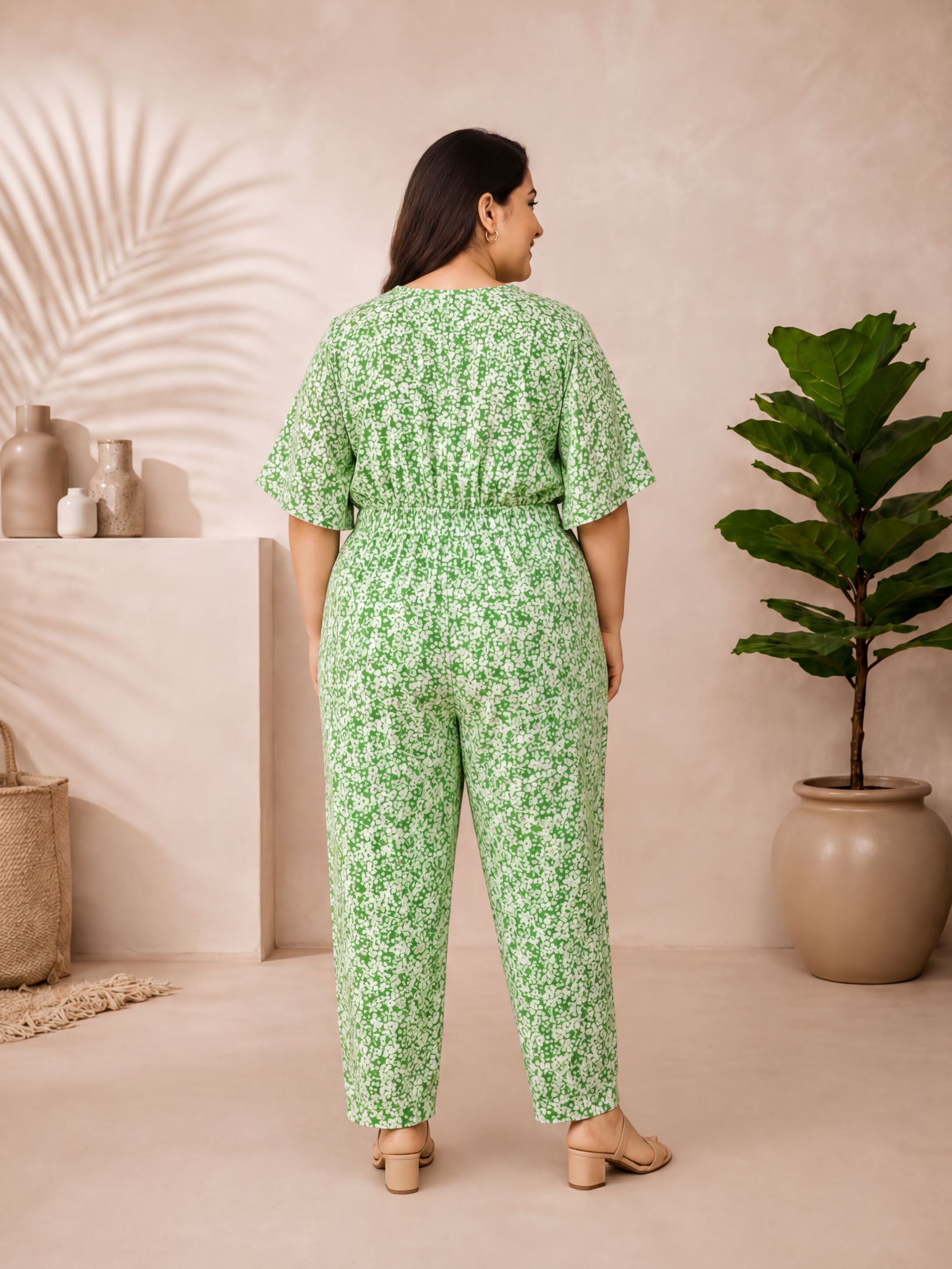 A woman wearing a green floral jumpsuit stands in a room with a plant and decorative items.