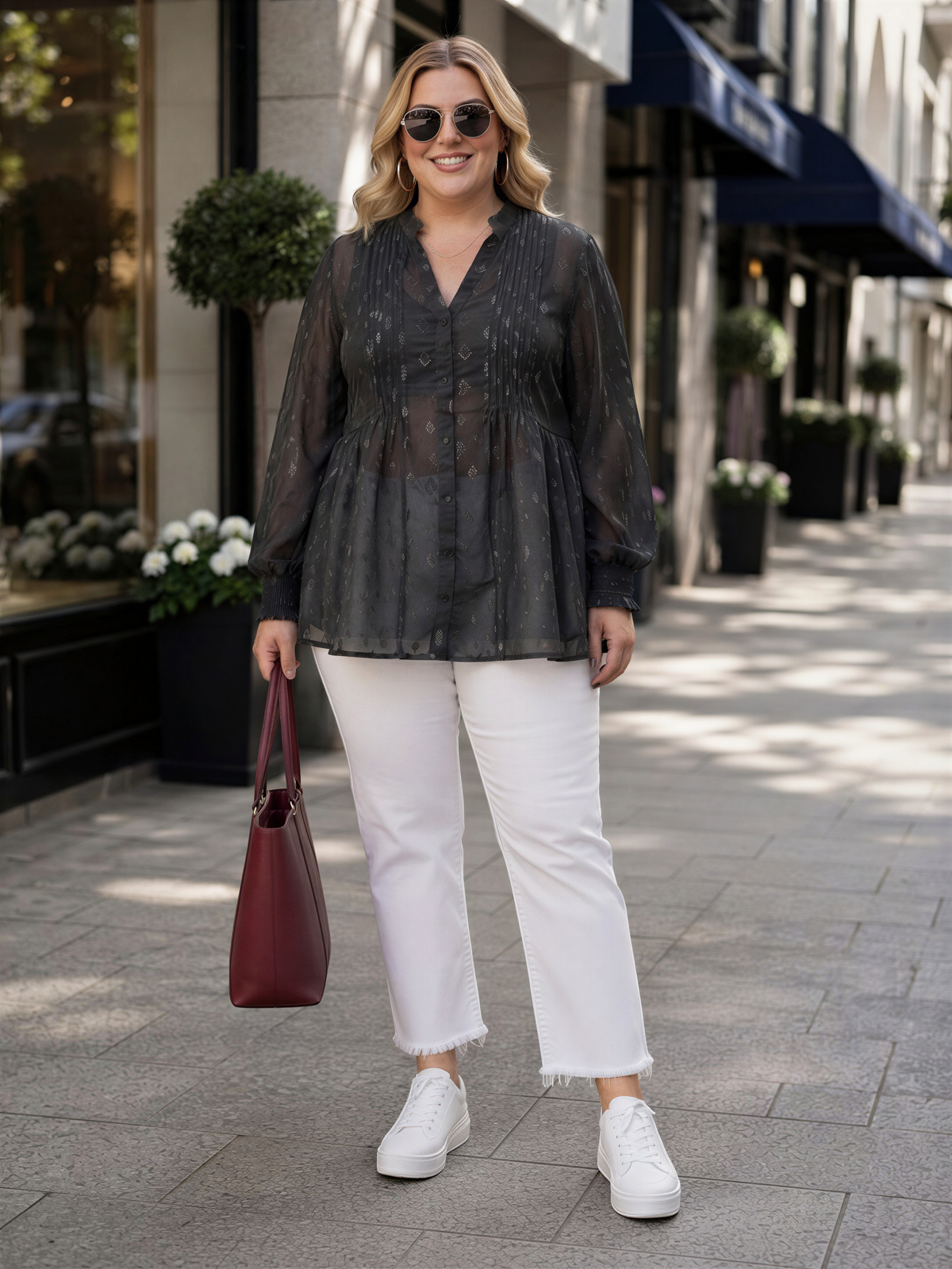 A woman wearing a black blouse, white pants, and white sneakers is standing on a sidewalk, smiling and holding a red handbag.
