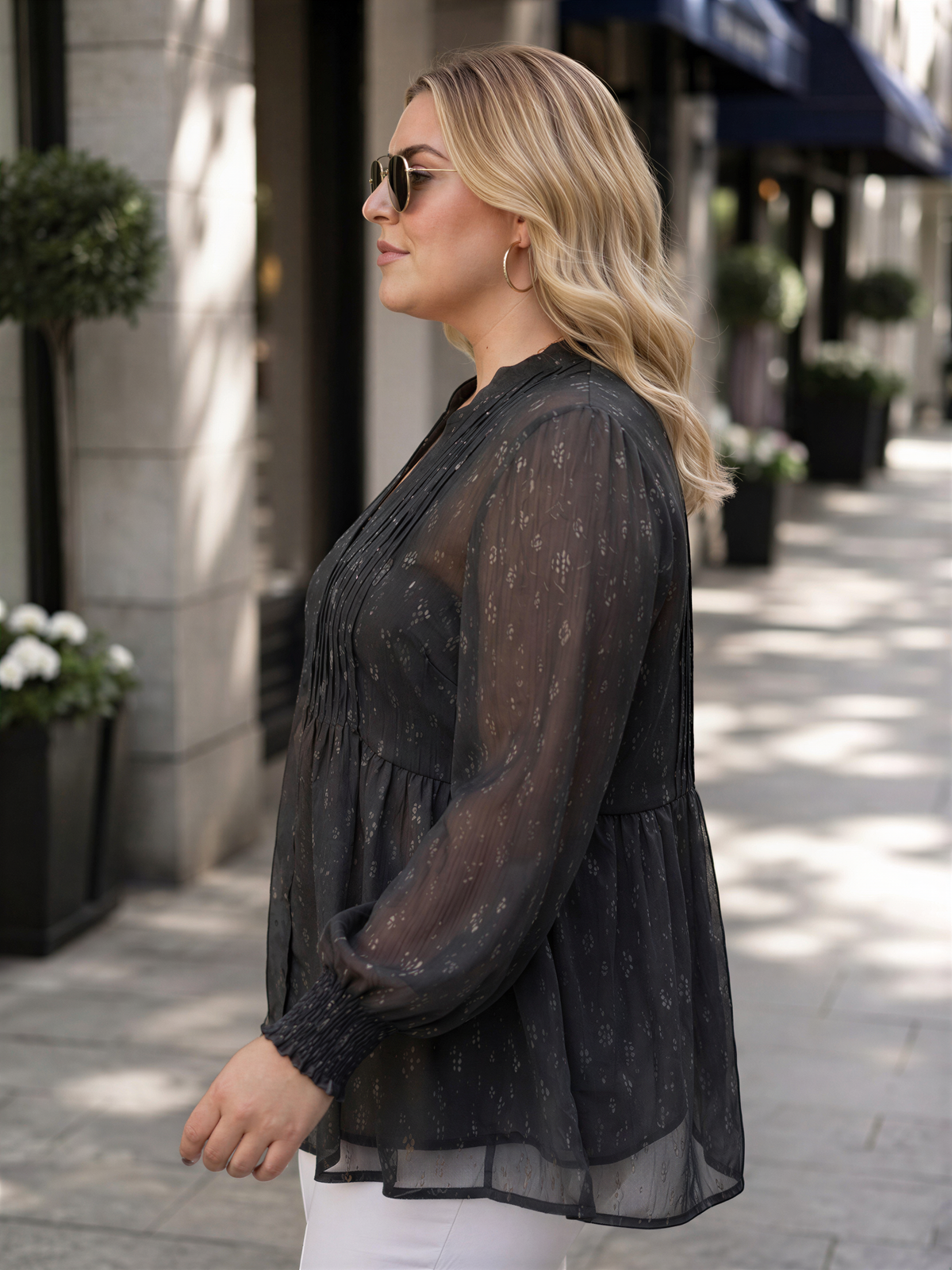 A woman wearing a black blouse with a patterned design, sunglasses, and white pants is standing on a sidewalk.