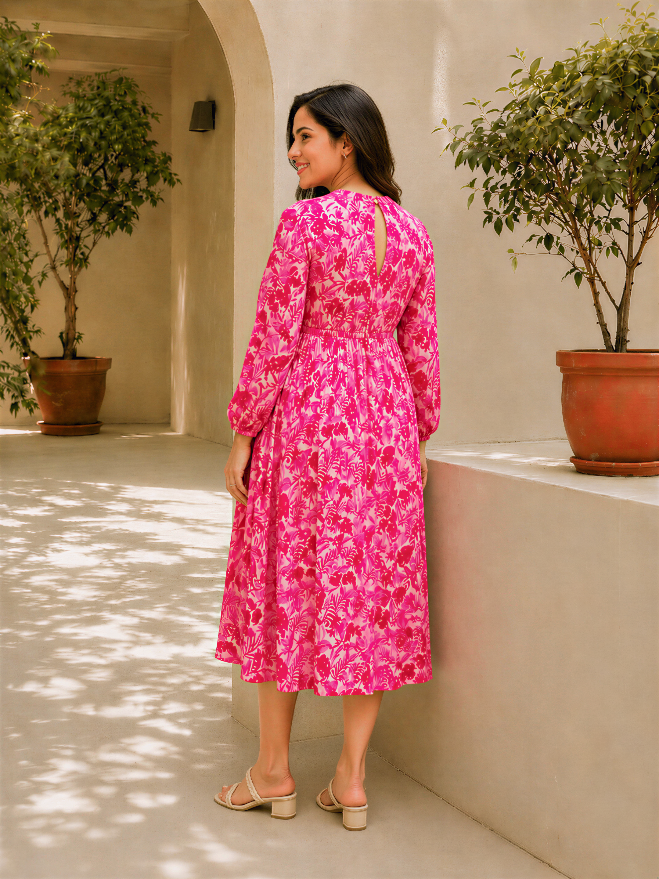A woman in a pink floral dress stands in a hallway with potted plants on either side, smiling at the camera.