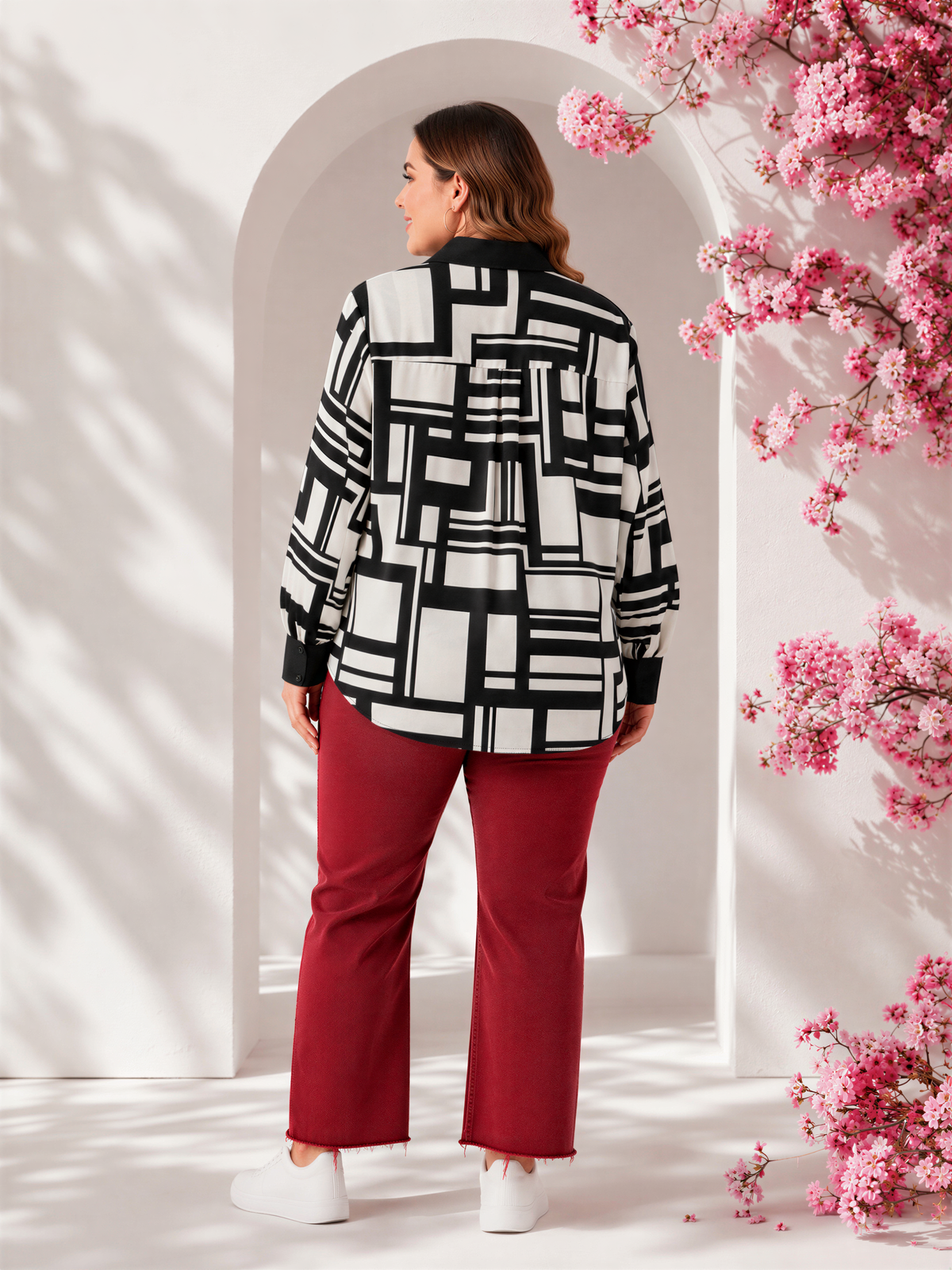 A woman wearing a black and white patterned shirt and red pants stands in front of a white archway with pink flowers.