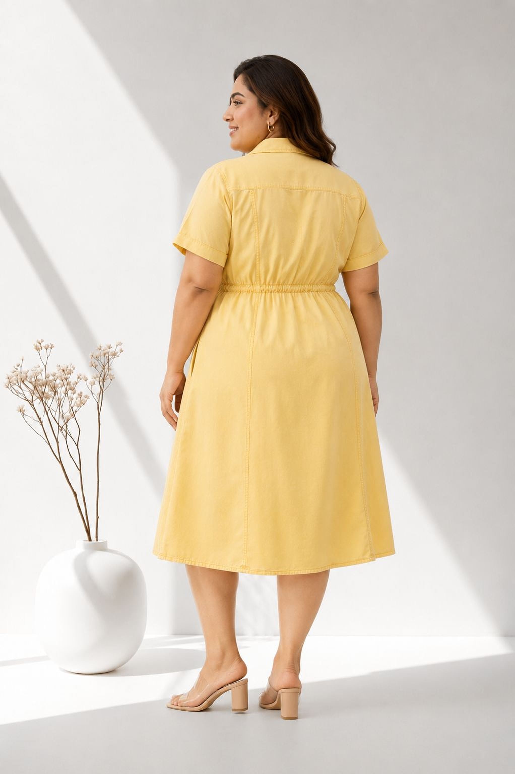 A woman wearing a yellow dress stands in front of a white vase with flowers, facing away from the camera.