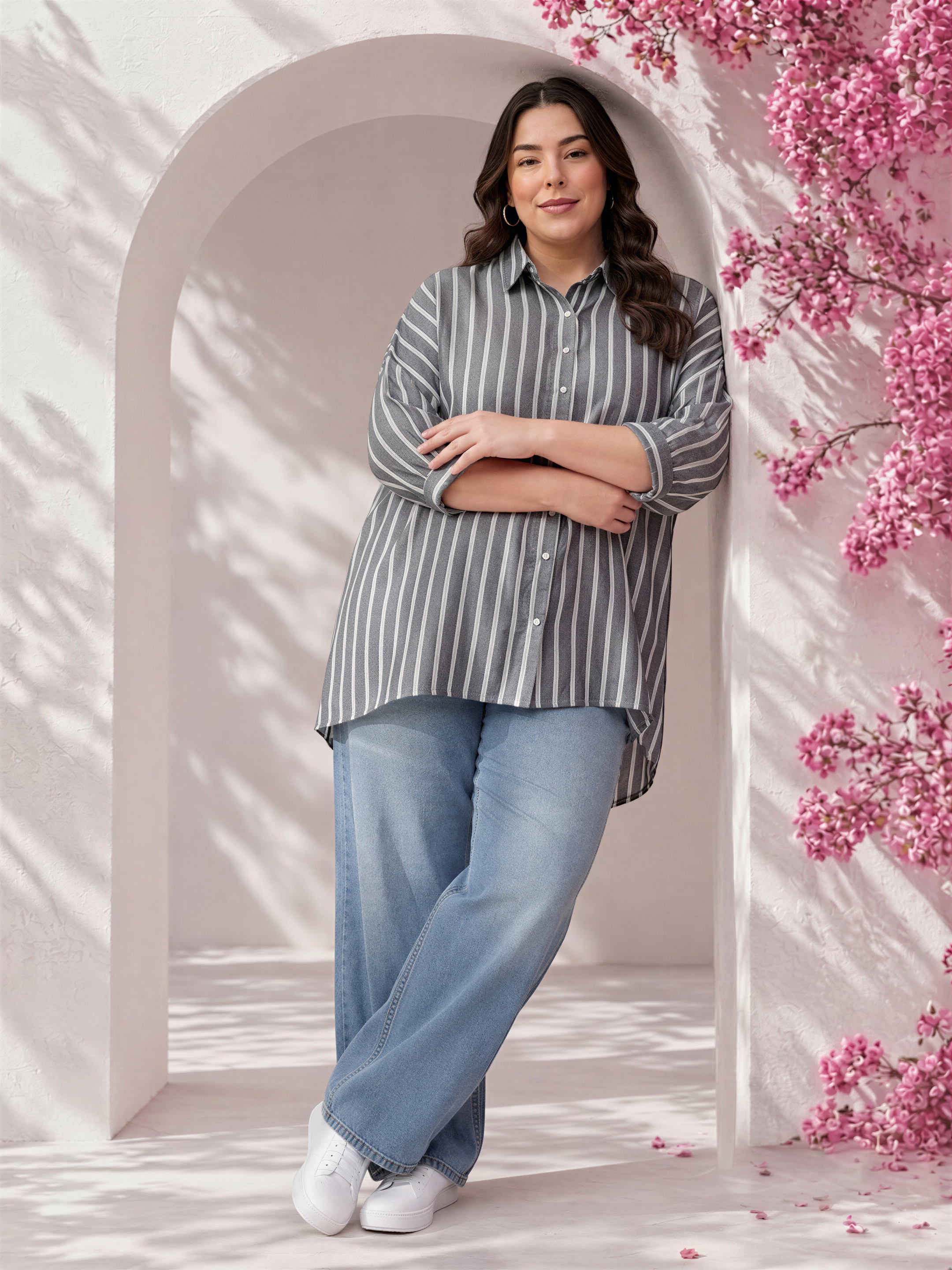 A woman in a striped shirt and jeans stands with her arms crossed in front of a pink flowering archway.