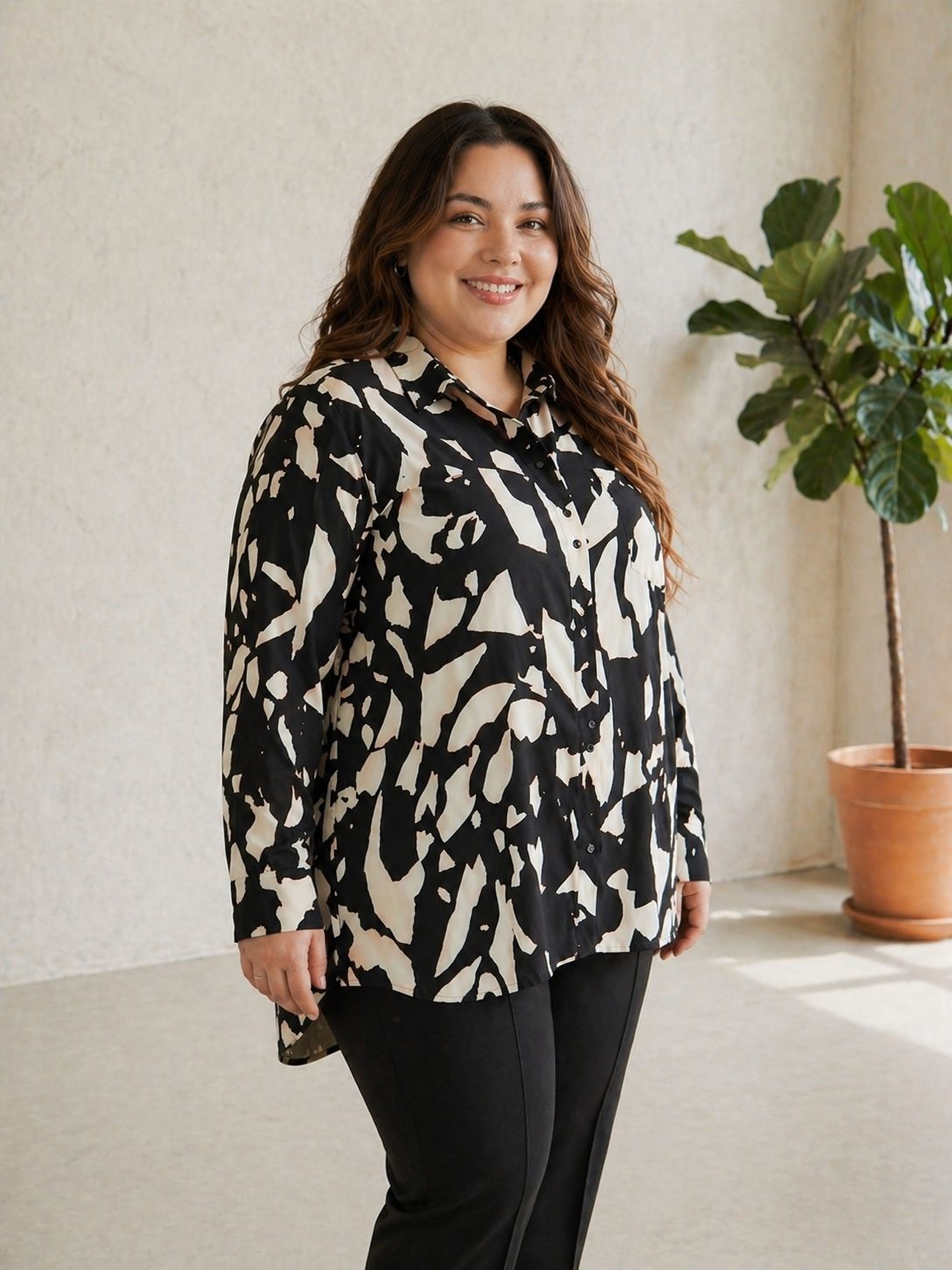 A woman with long brown hair is standing in a room, smiling at the camera. She is wearing a black and white patterned blouse and black pants. There is a potted plant in the background.