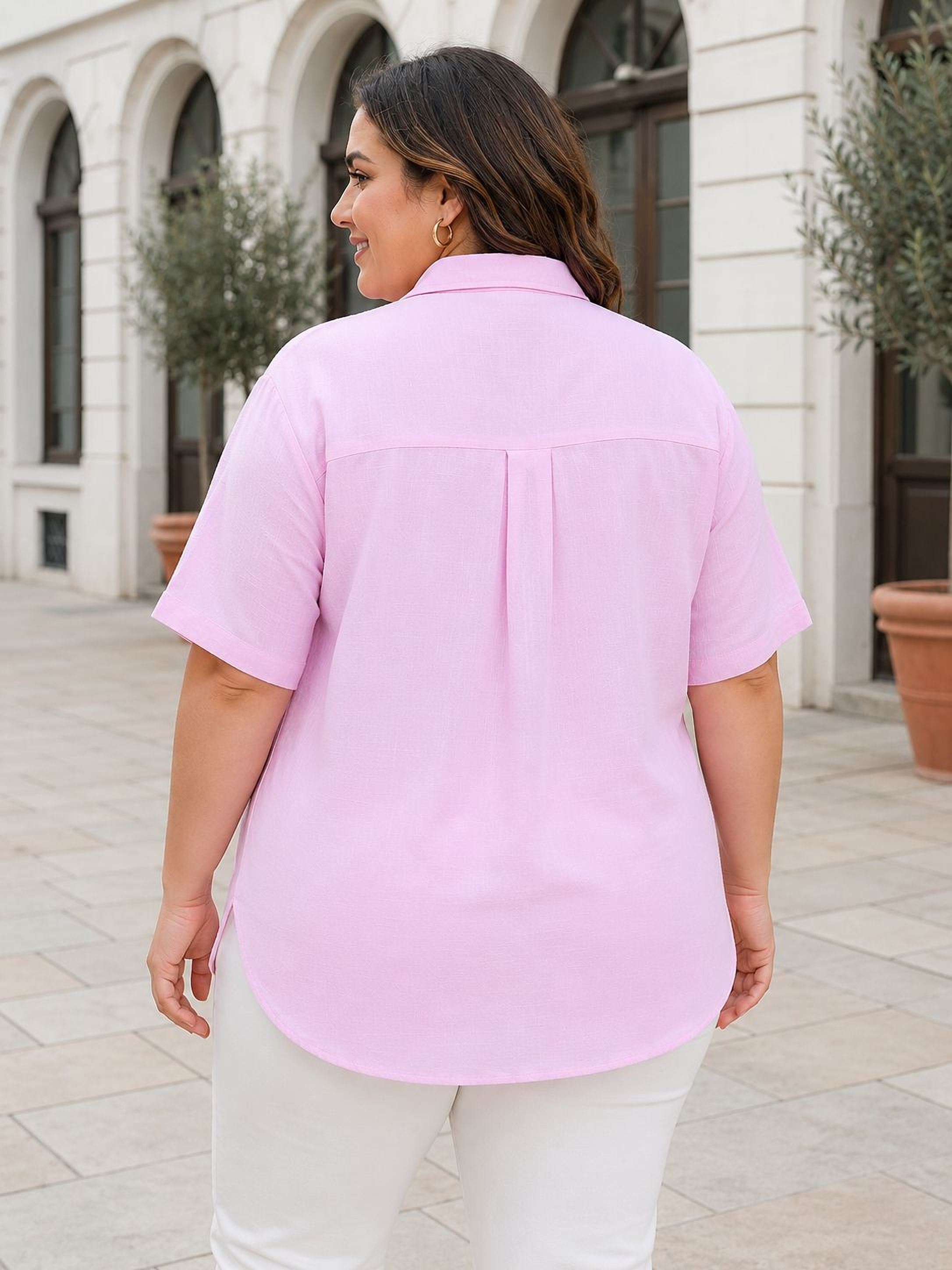 A woman wearing a pink shirt and white pants is standing in front of a building with arched windows.