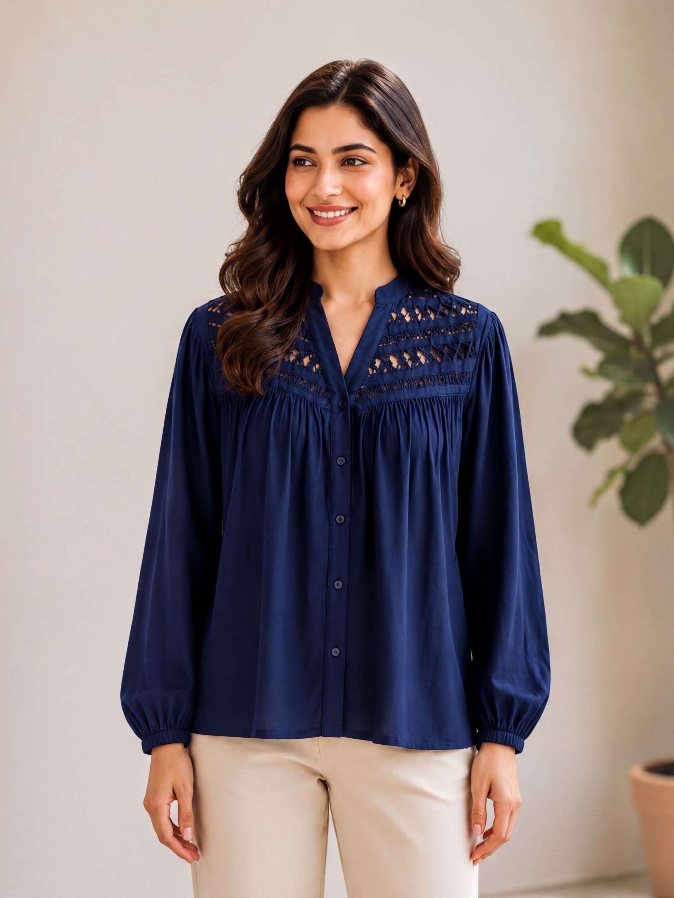 A woman with long brown hair wearing a navy blue blouse with lace detailing and puff sleeves stands in front of a white wall, smiling at the camera.