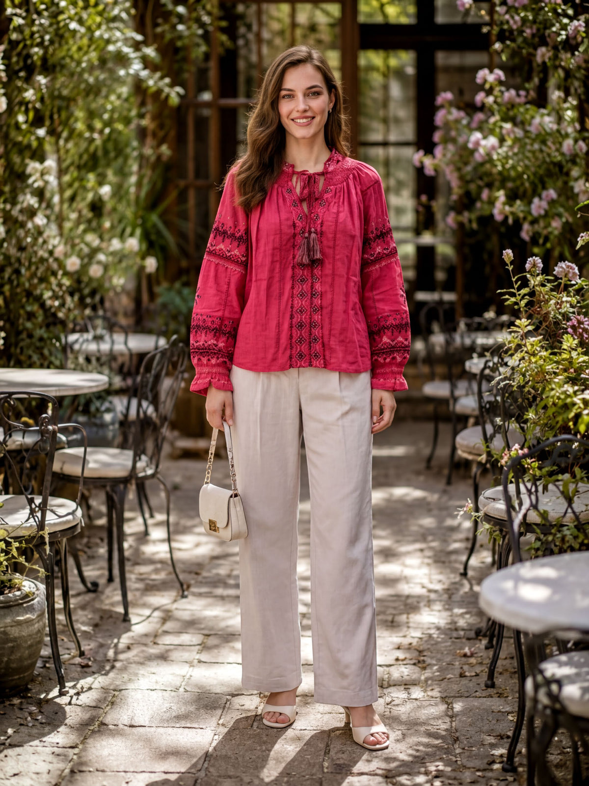 A woman wearing a red blouse with embroidered patterns and beige pants stands in a garden setting with tables and chairs.