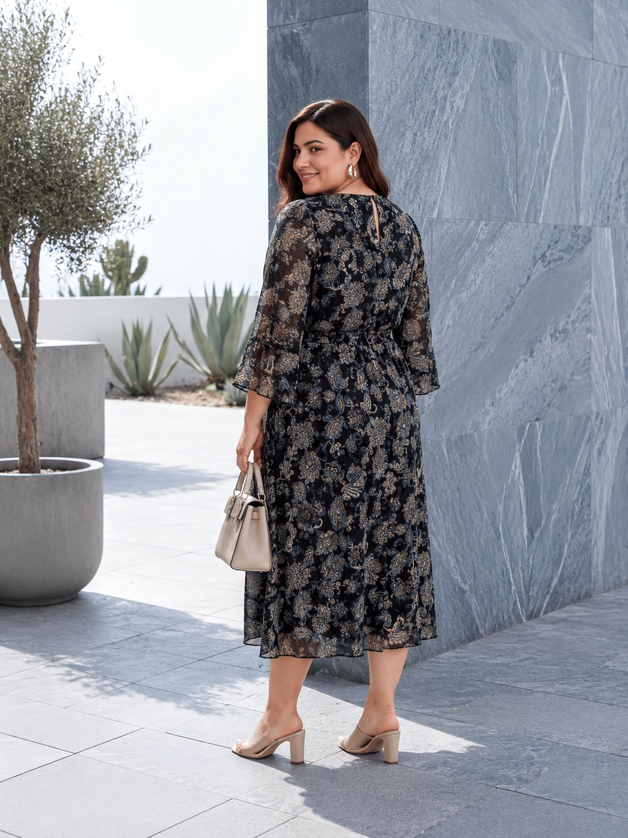 A woman wearing a black and white floral dress stands in front of a gray wall, smiling and holding a beige handbag.